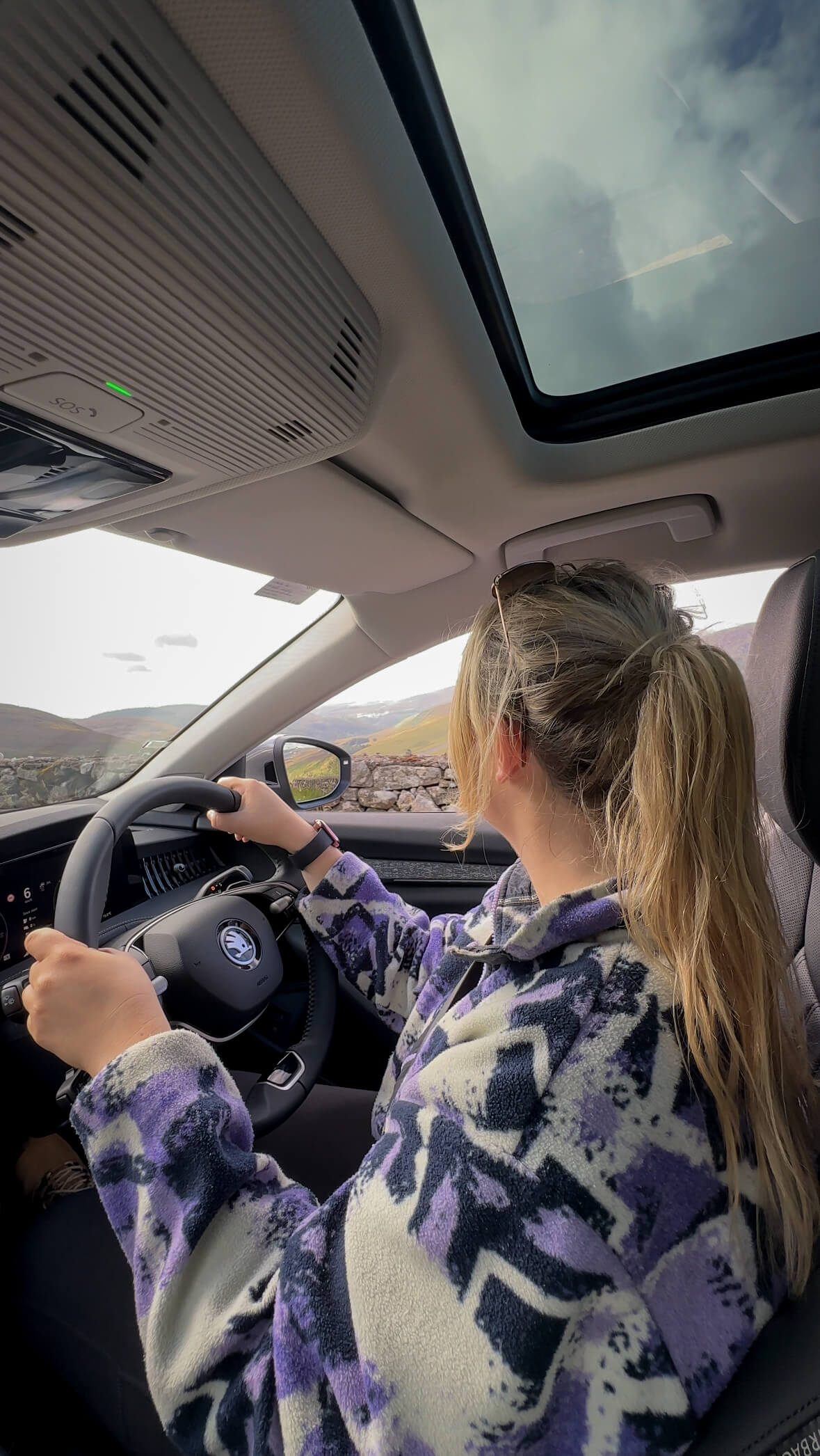 Woman driving on scenic Irish road with rolling hills and stone walls