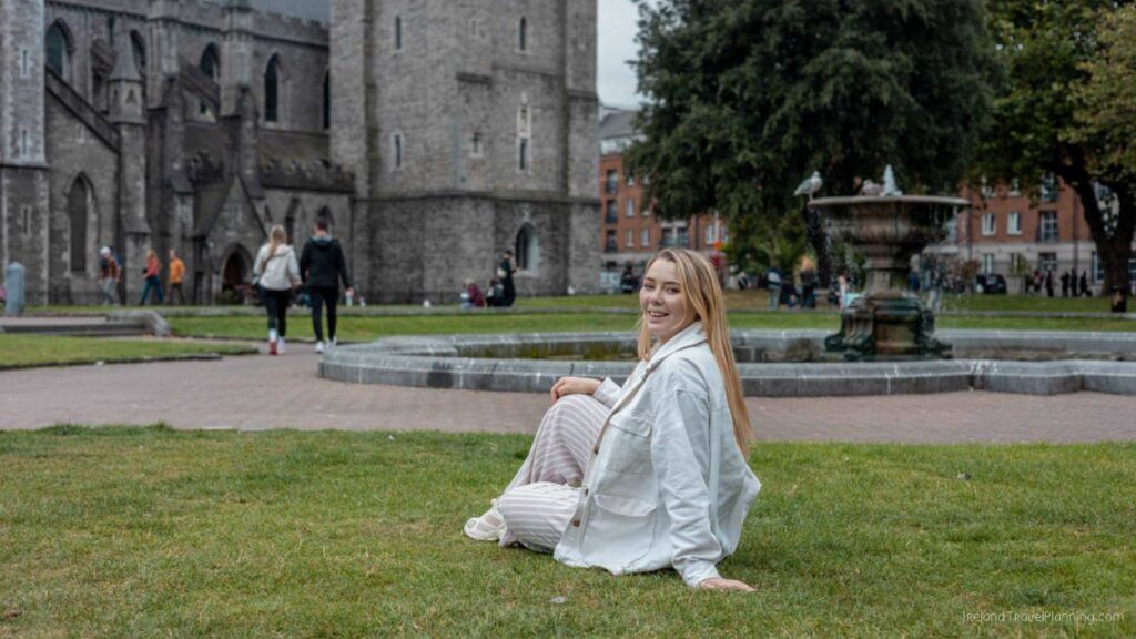Woman sits in St. Patrick's Park, a free thing to do in Dublin, with the cathedral and fountain in the background.