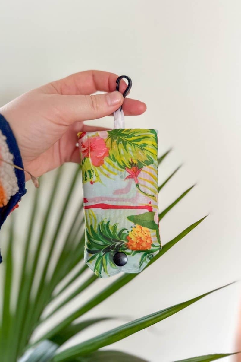Compact, folded reusable shopping bag with a tropical print, held by a hand.