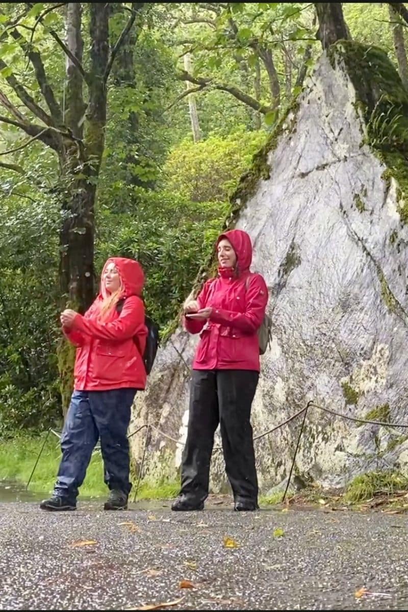 Two women in red raincoats stand near a large rock in a forest, offering Ireland travel tips.