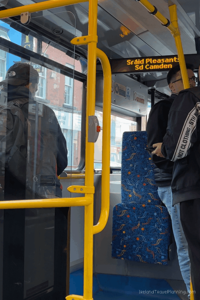 Inside a Dublin bus, passengers stand near yellow poles. Destination sign shows "Srรกid Pleasant, Camden.