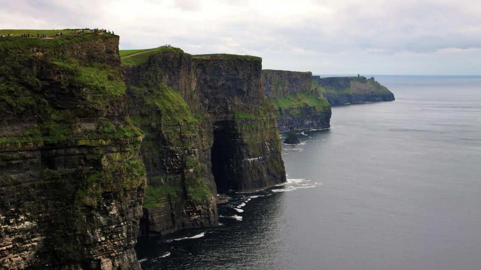 Dramatic view of the Cliffs of Moher in Ireland, with green grass on top and a sea cave.