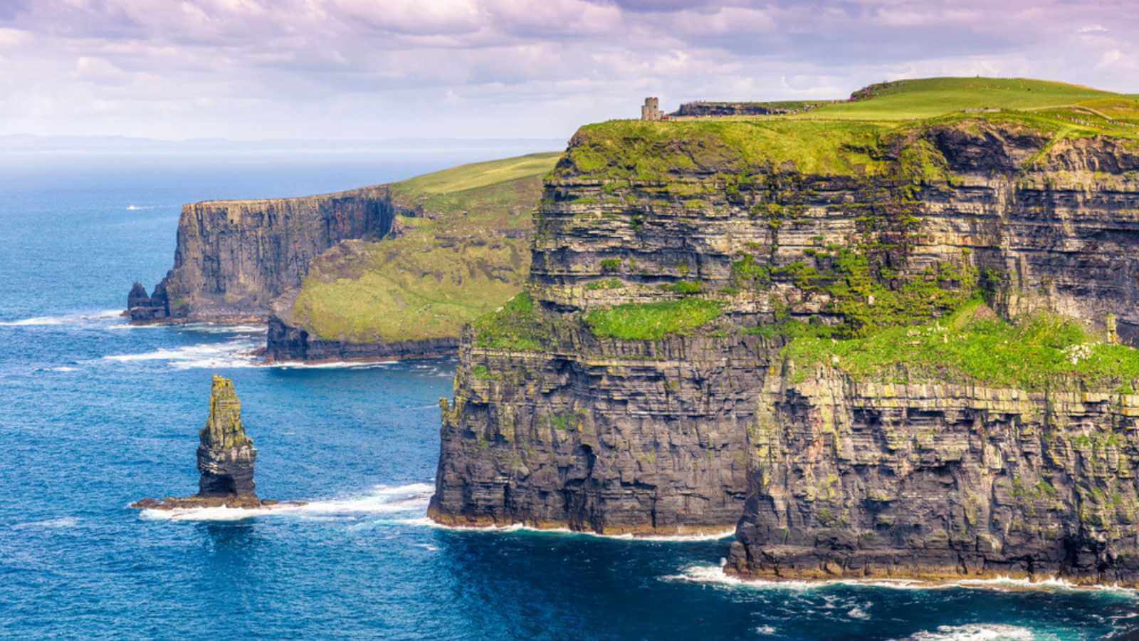 Dramatic Cliffs of Moher, Ireland, with green grass on top and a sea stack in the blue ocean.