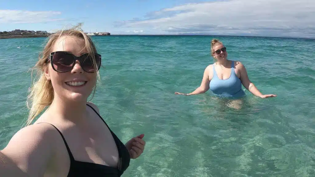 Two women enjoying the clear turquoise water at a sunny beach, smiling at the camera.
