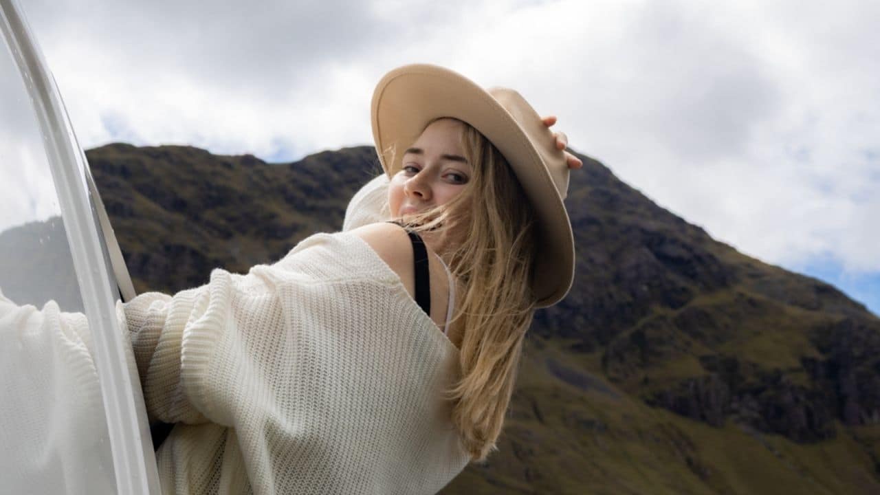 Woman in hat leans out of camper van window, mountain backdrop.