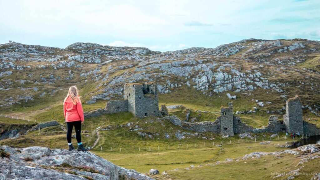 Woman views ruins of Three Castle Head in Ireland. Stone structure, green grass, rocky hills.