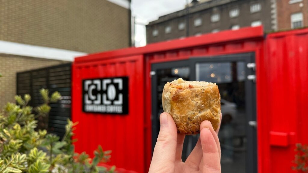 Sausage roll held in front of a red container cafe, "Brewbird Coffee" sign visible.