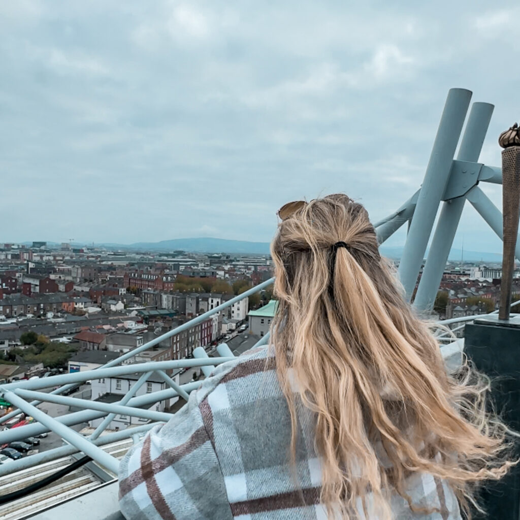 Woman with blonde hair looking out over Dublin from a high vantage point. Cityscape under cloudy sky.