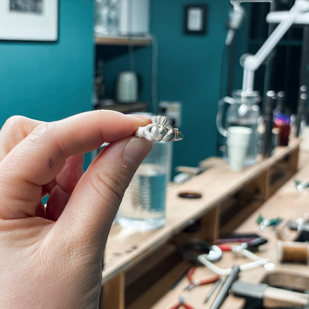 Hand holding a silver Claddagh ring, jewelry making tools in the background.