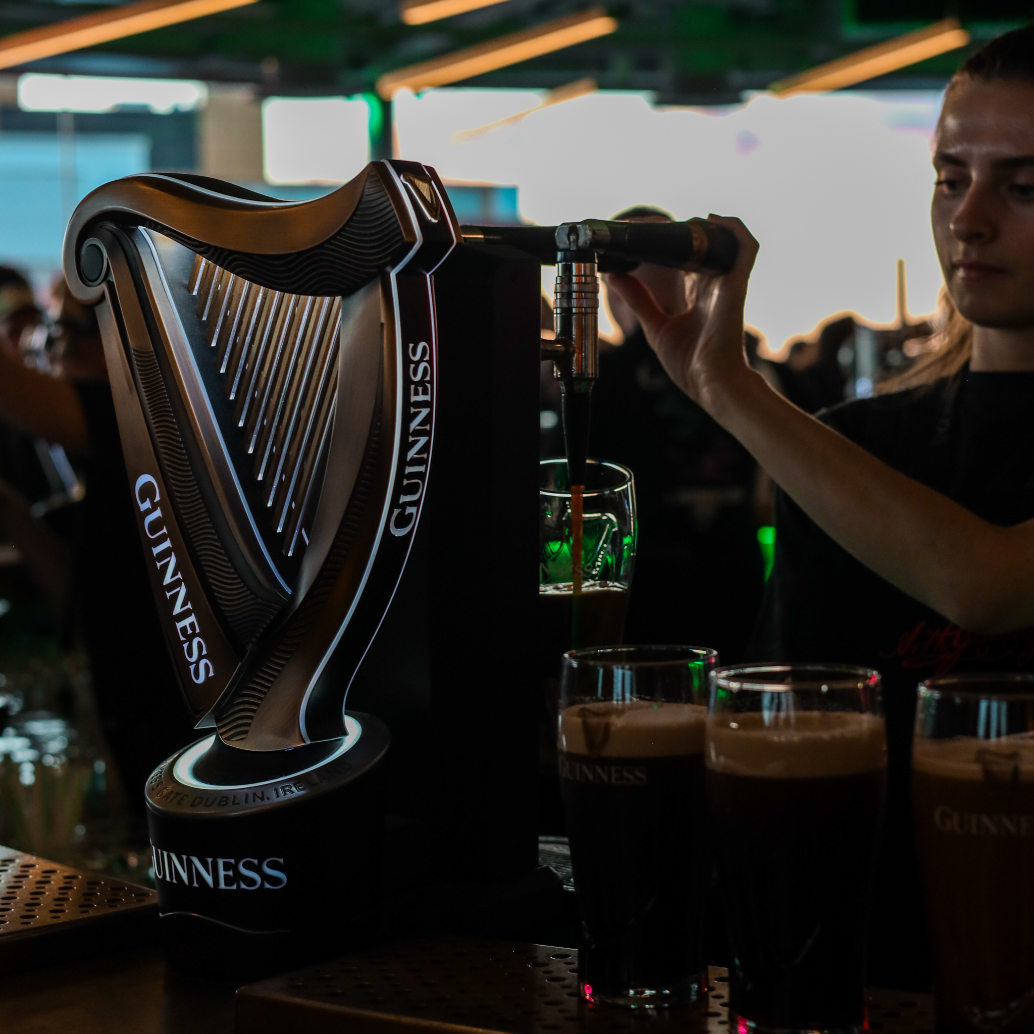 Guinness being poured from a tap at a bar, with other pints ready. "Guinness" is visible on the tap and glasses.