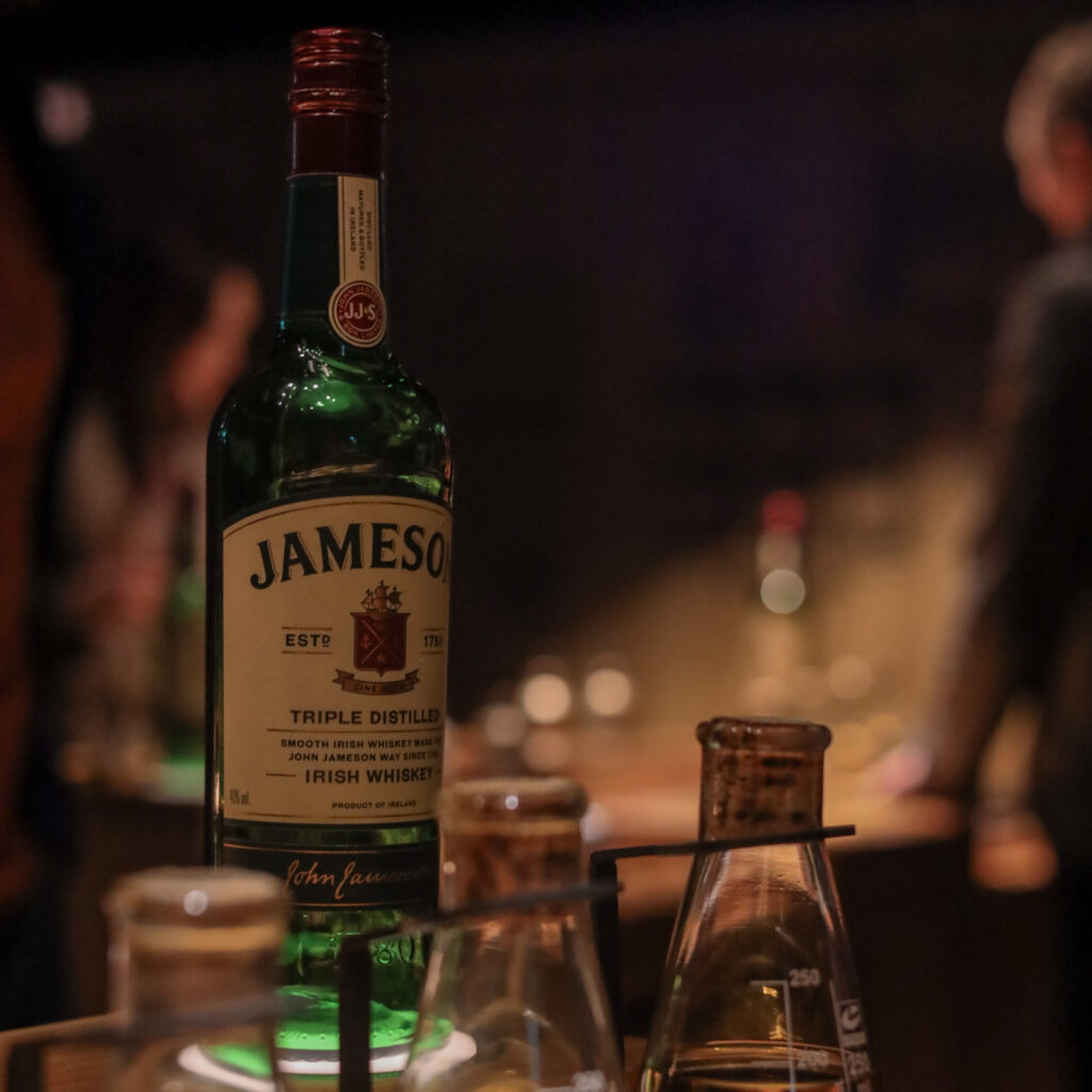 Jameson Irish Whiskey bottle with lab beakers in background. "Triple Distilled Irish Whiskey" text visible.