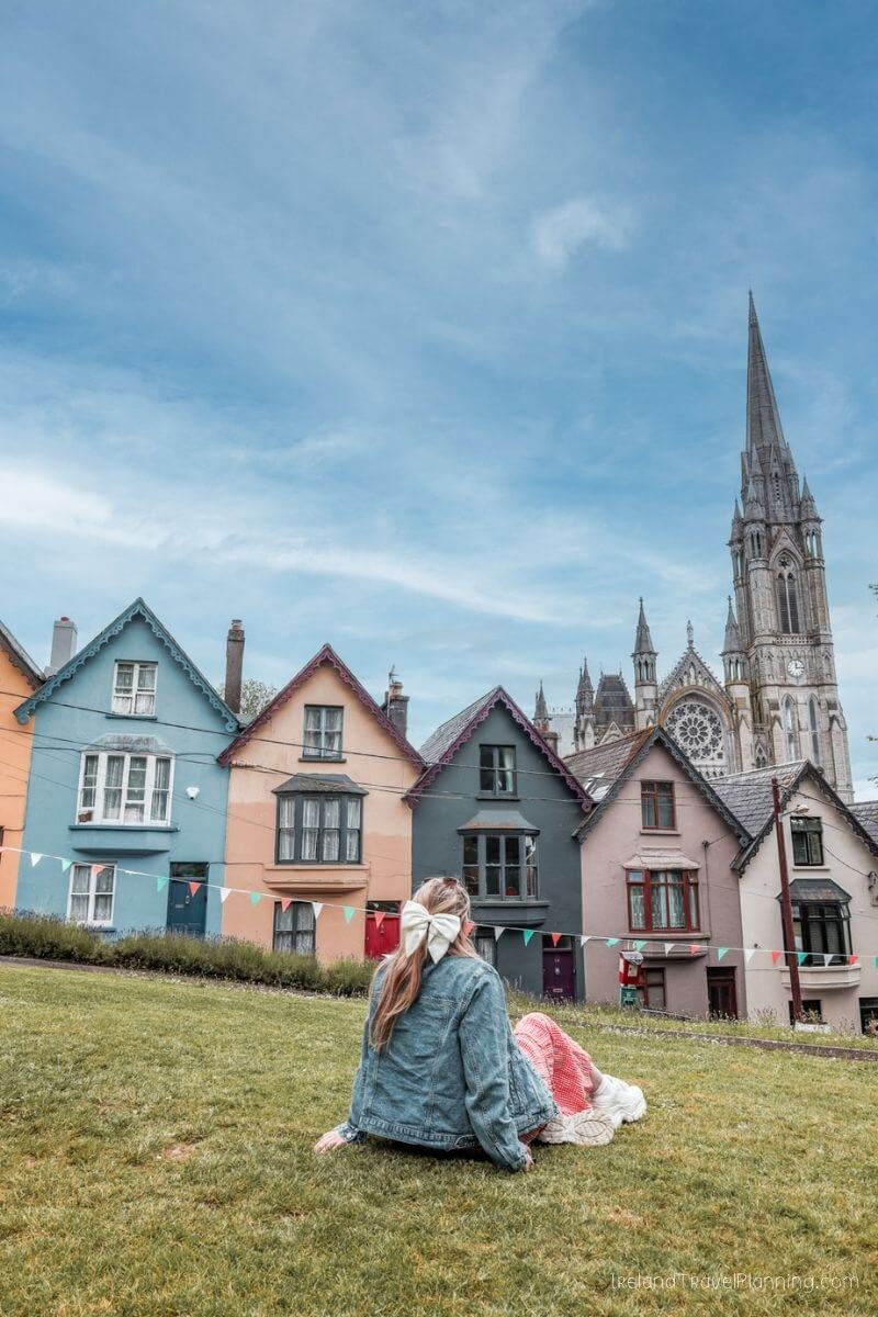 Woman in denim jacket sitting on grass, colorful houses in Cork City behind