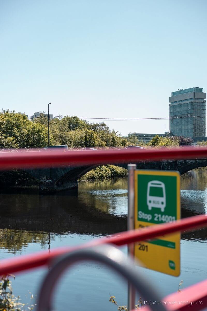 Bus stop sign near a river, with a bridge and city buildings in the distance