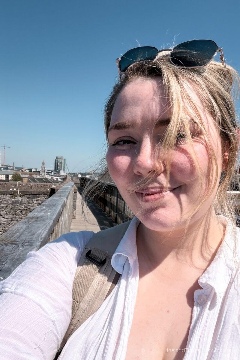 Blonde woman smiling in Cork City, wearing sunglasses, with city skyline in background