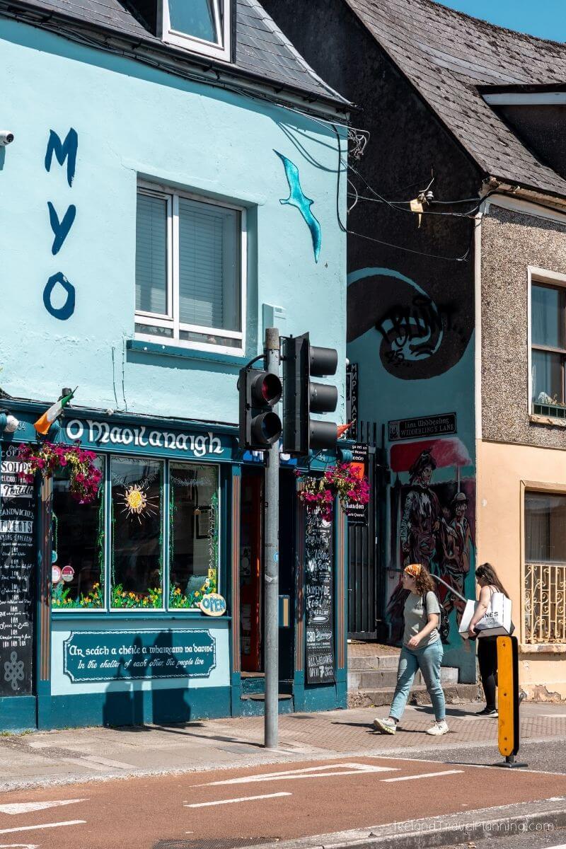 Two women walking past a blue building with Irish text and murals in Cork City