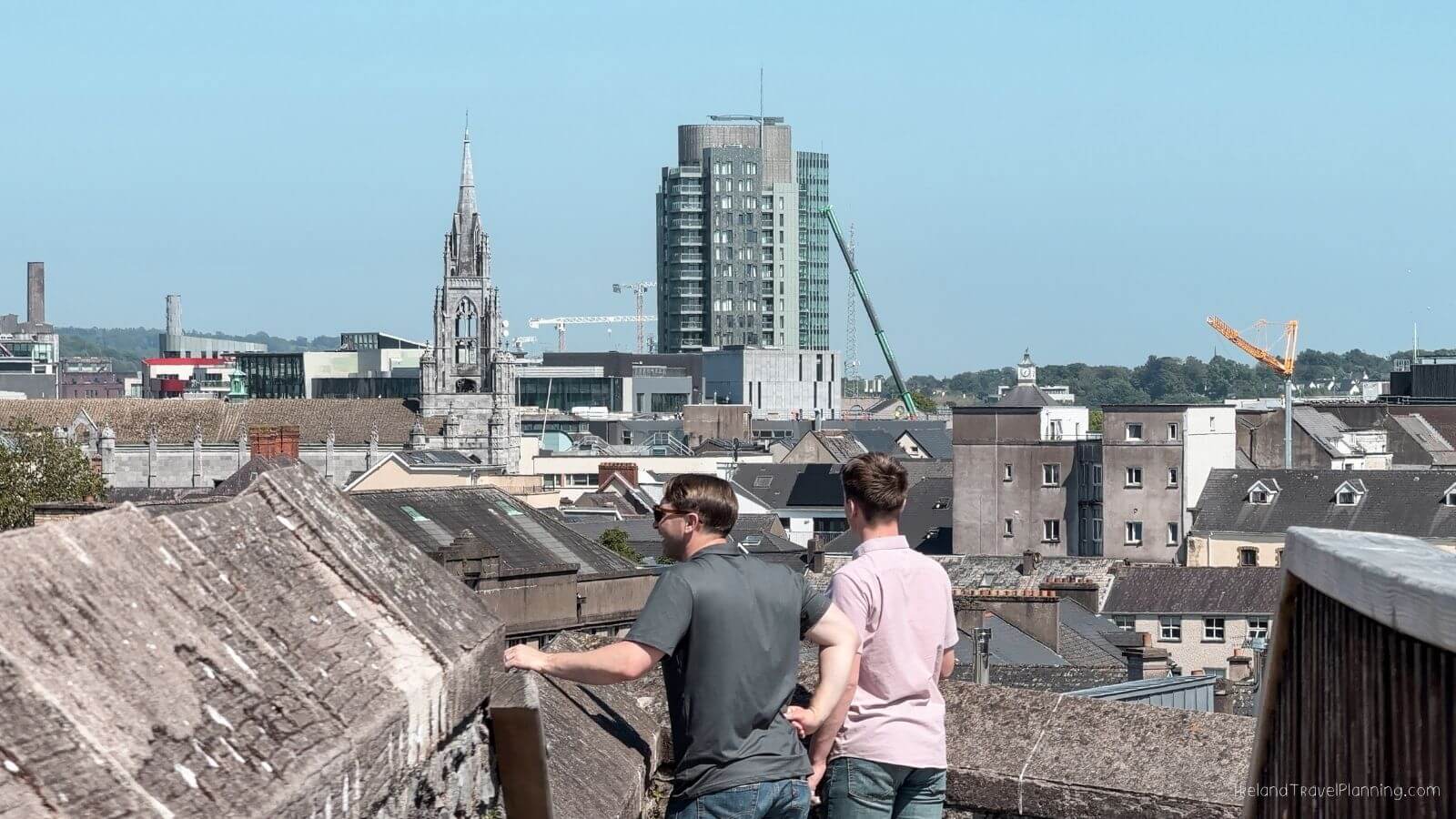 Two men overlook Cork City rooftops with historic and modern buildings.