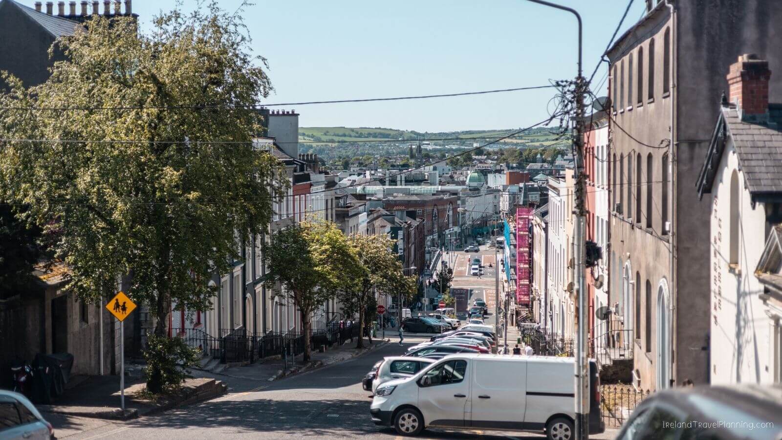 View down a steep street in Cork City, Ireland, with colorful buildings and parked cars.