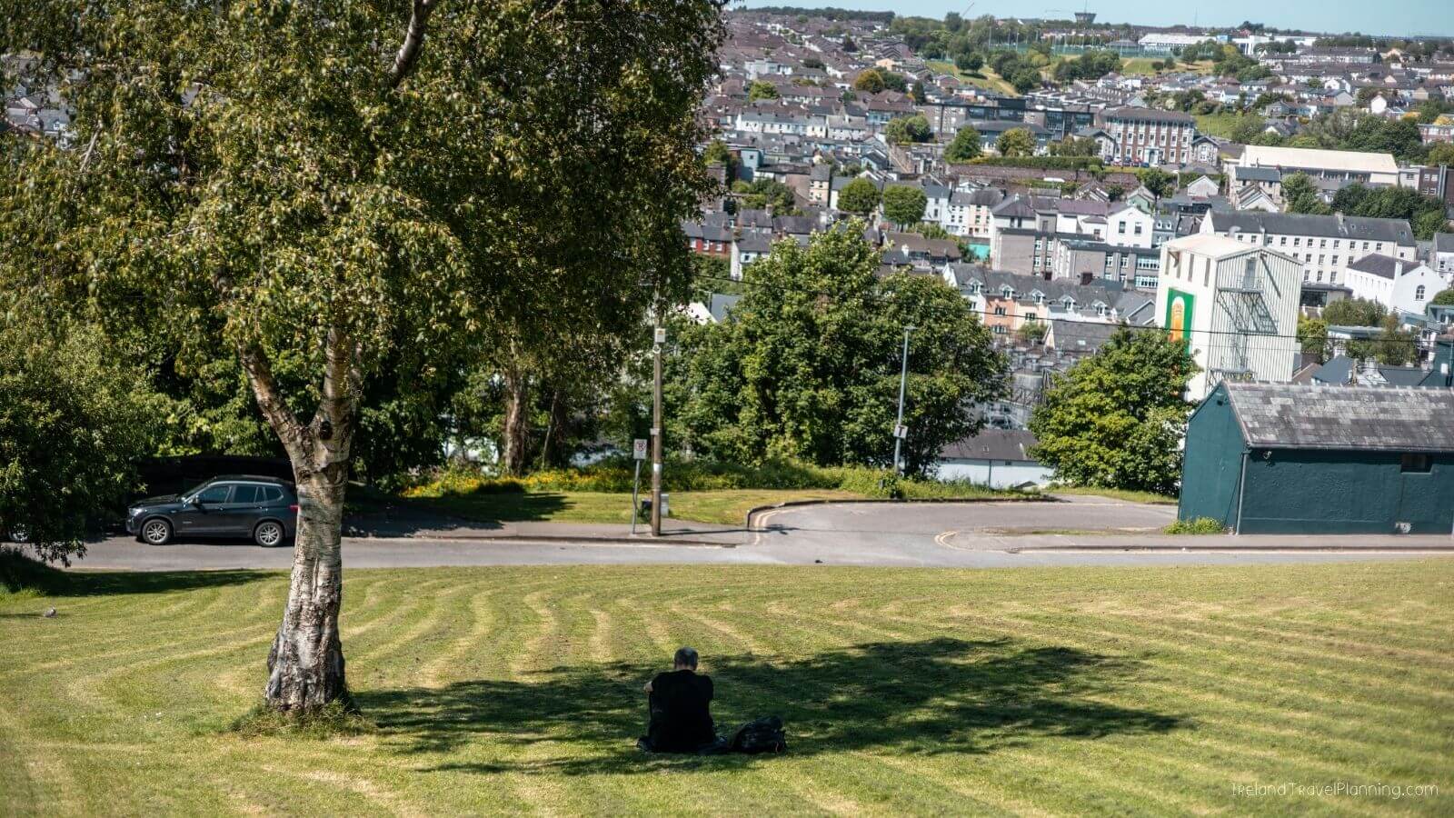 A man sitting on the grass under a tree, overlooking Cork City