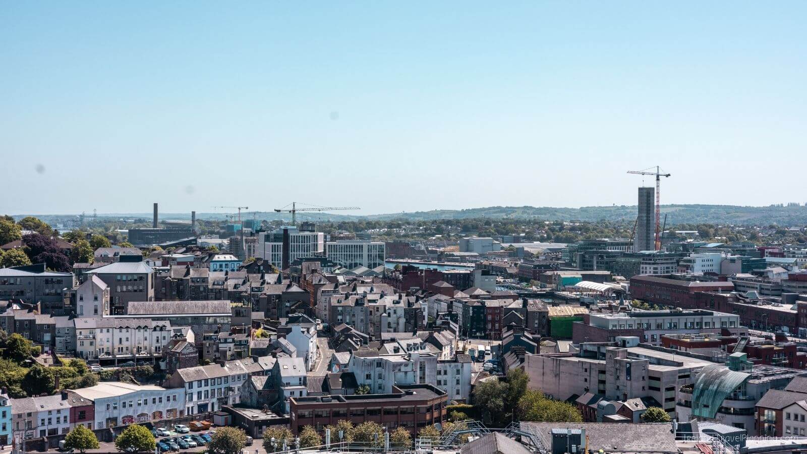 Aerial view of Cork City with buildings, cranes, and trees under a blue sky