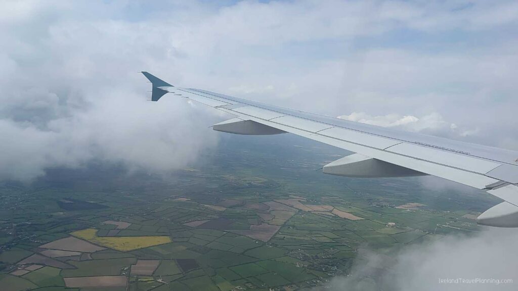 Airplane wing over patchwork green fields and clouds, Ireland travel view.
