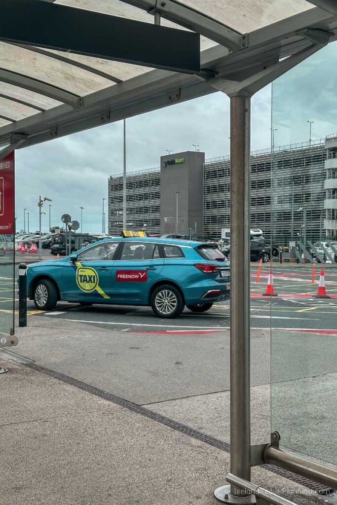 Blue taxi with "TAXI" and "FREE NOW" logos waiting at an airport bus stop.