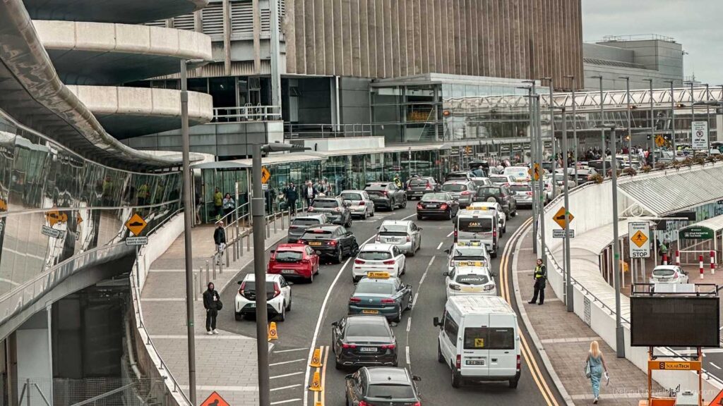 Busy airport arrivals area with cars, taxis, and people walking.
