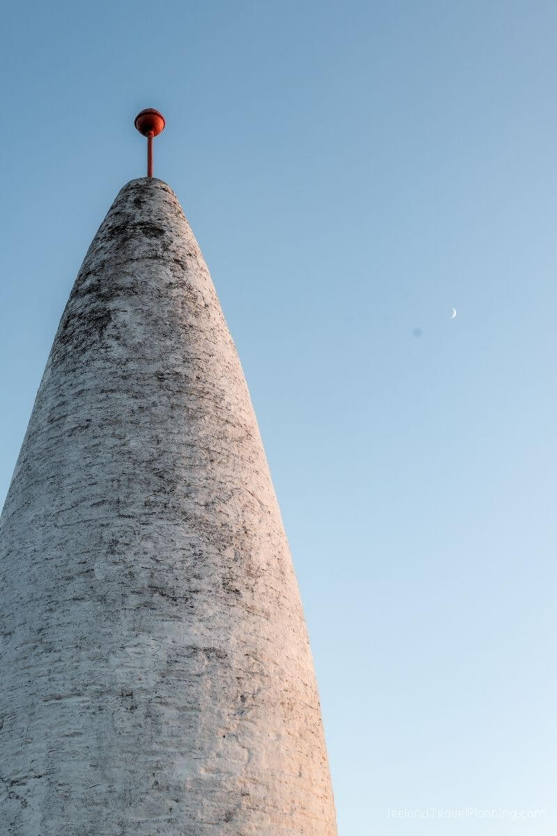 Conical lighthouse with red top against a clear blue sky and crescent moon