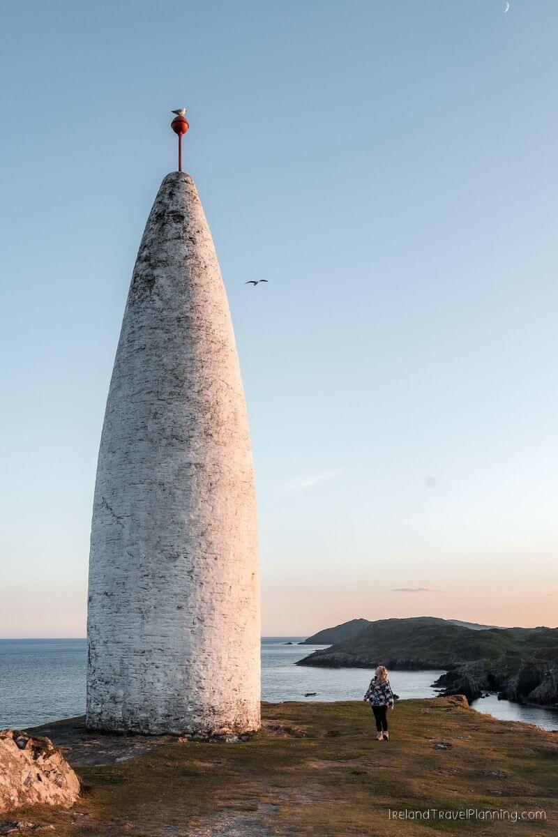 Cone-shaped marker on a grassy cliff overlooking the sea, with a woman walking nearby.