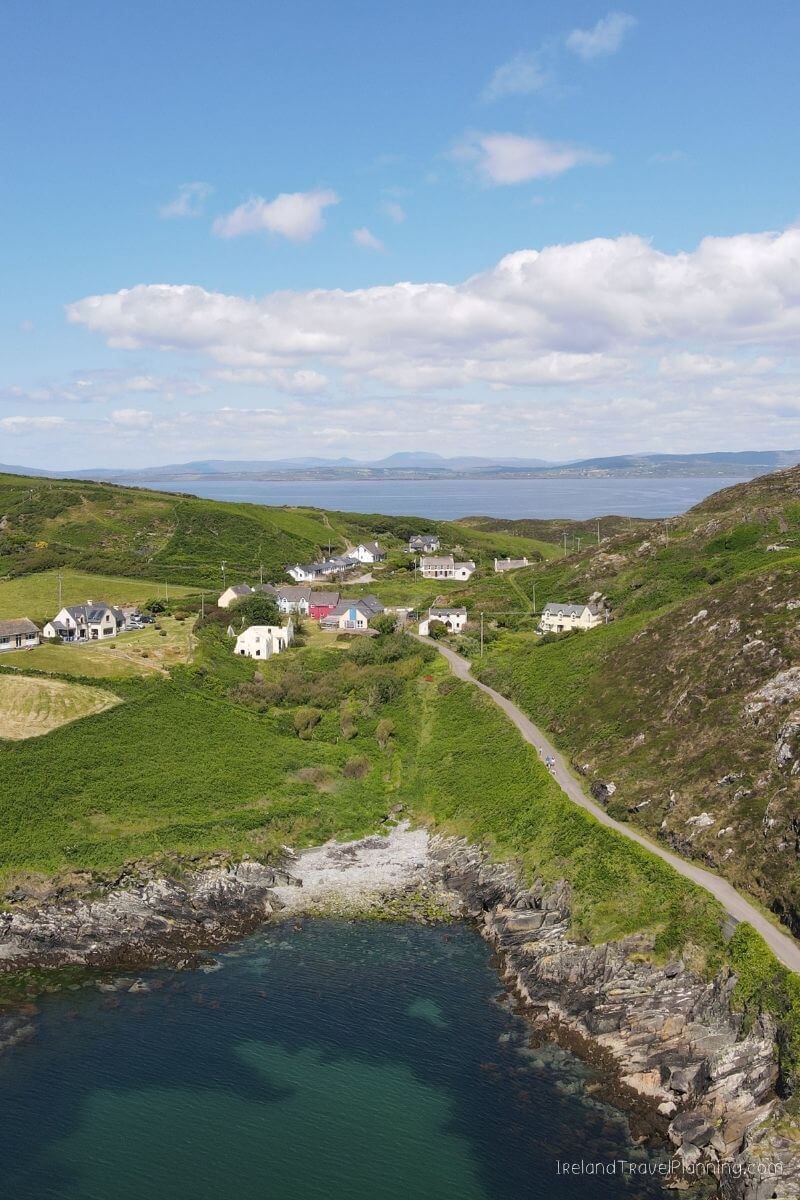 Aerial view of a coastal Irish village with green fields, houses, and a path by the sea