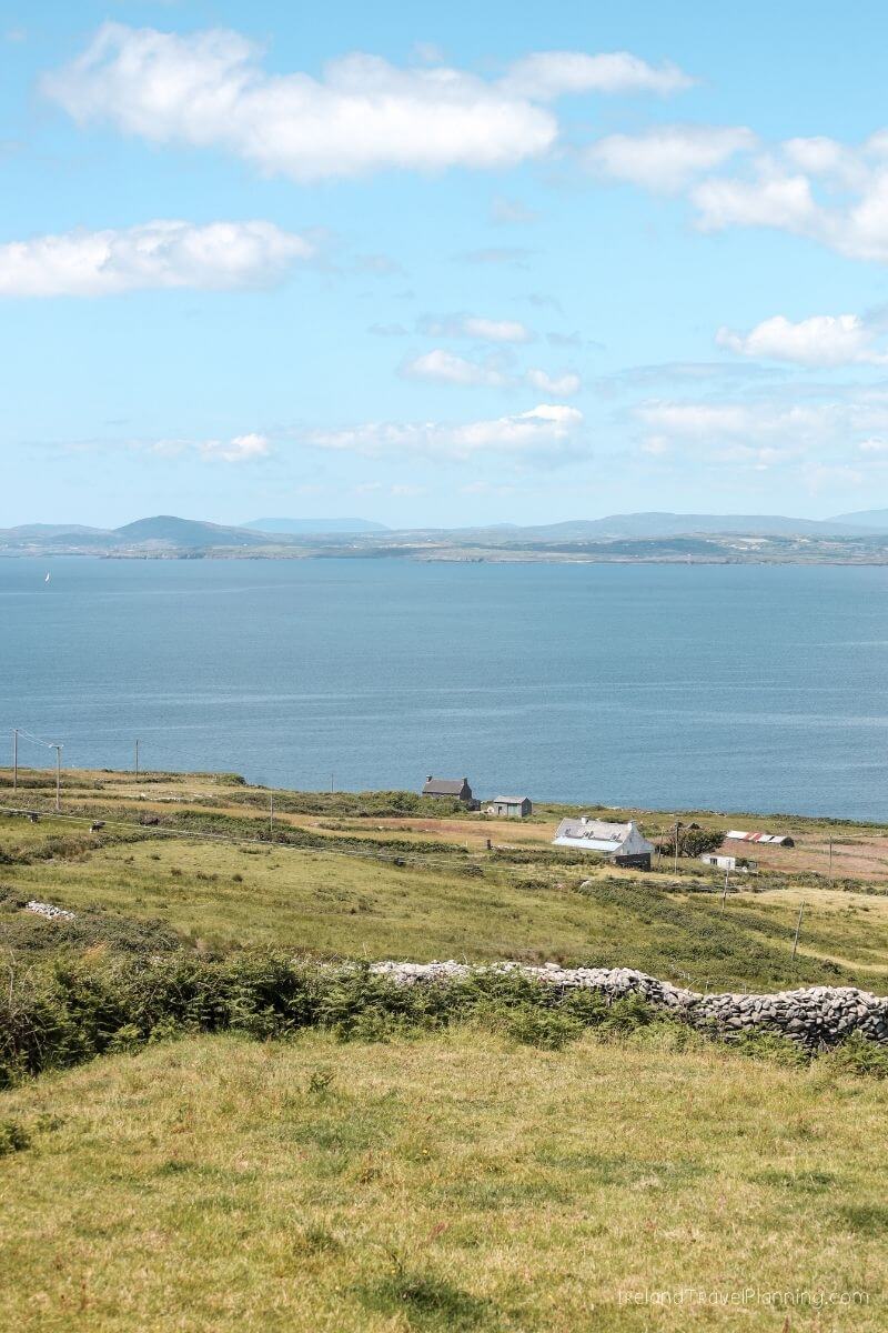 Coastal Ireland landscape with rolling green hills, a calm blue sea, and scattered farmhouses.