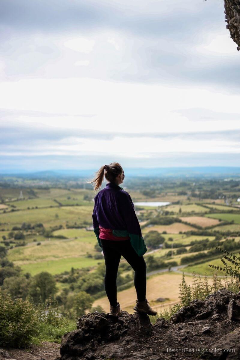 Woman looking over lush green Irish landscape from a rocky viewpoint.