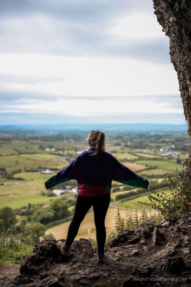 Woman in colorful jacket overlooking rolling green hills from a cave in Ireland