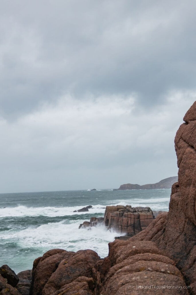 Rugged Irish coastline with crashing waves against red rocks under a cloudy sky.