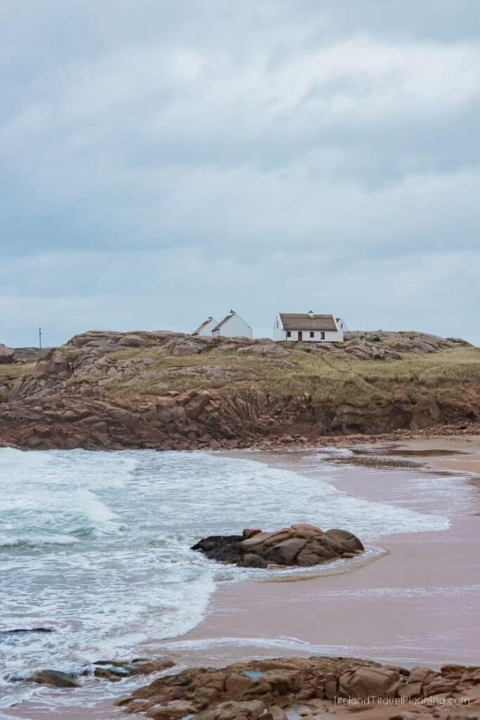 Two white cottages on a rocky, grassy coastline with waves crashing on a sandy beach.