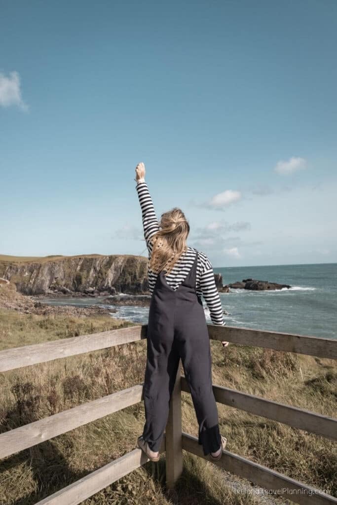Woman wearing striped shirt and overalls standing on wooden fence, looking at ocean