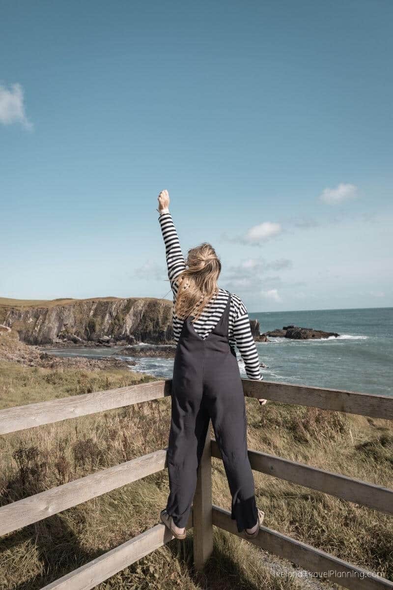 Woman wearing striped shirt and overalls standing on wooden fence, looking at ocean