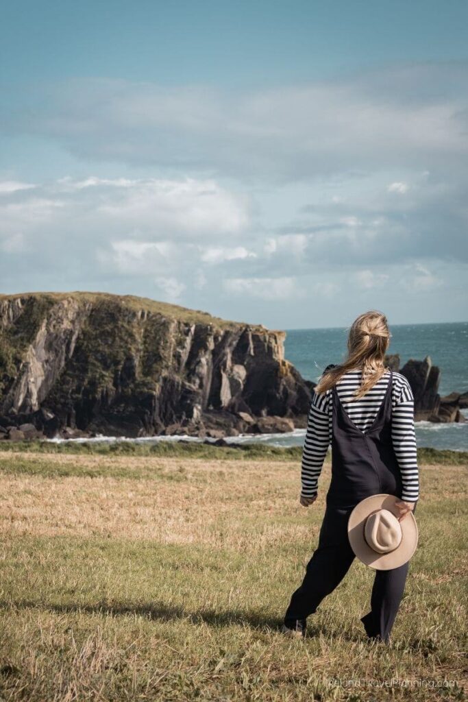 Woman in overalls and hat overlooks rugged coastline and sea