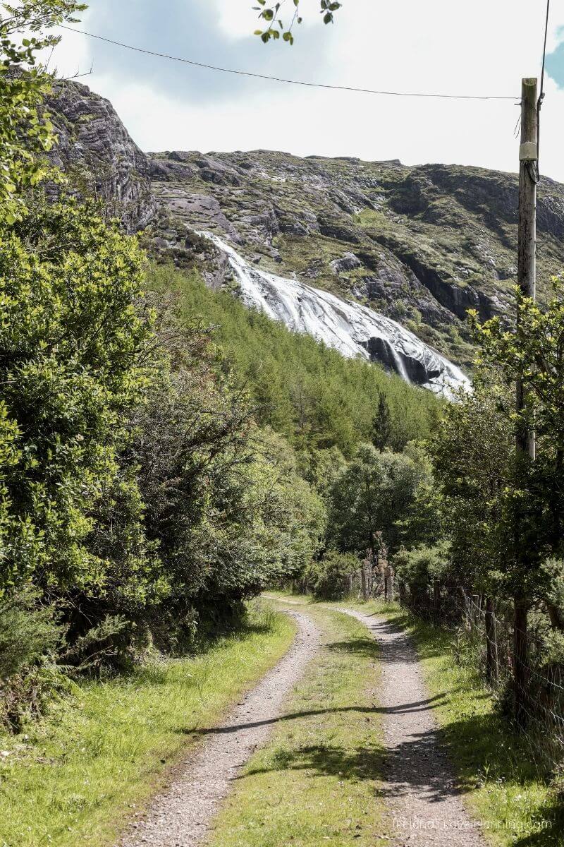 Scenic Irish waterfall cascading down a rocky mountain, with lush green trees and a dirt path.