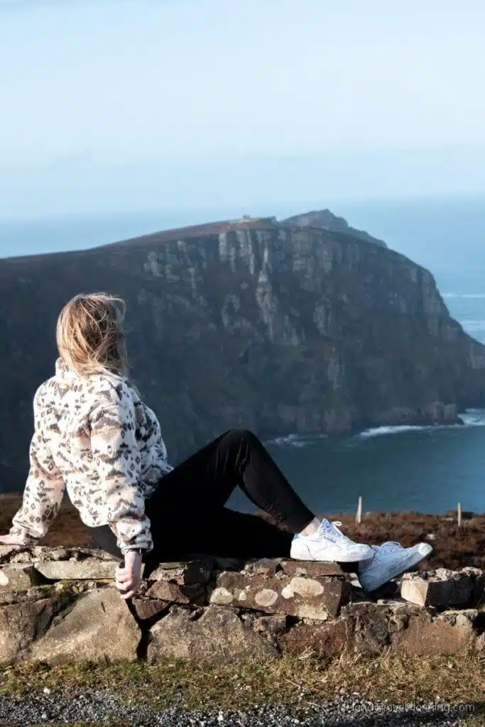 Woman sitting on a stone wall overlooking a rugged coastline and the ocean.
