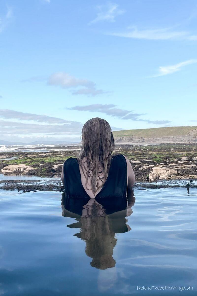 Woman in a natural ocean pool at the coast of Ireland, with waves and green cliffs in the background.