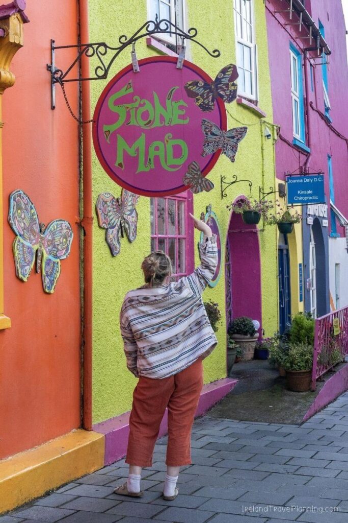 Woman points at colorful "Stone Mad" shop sign with butterflies in Kinsale, Ireland.