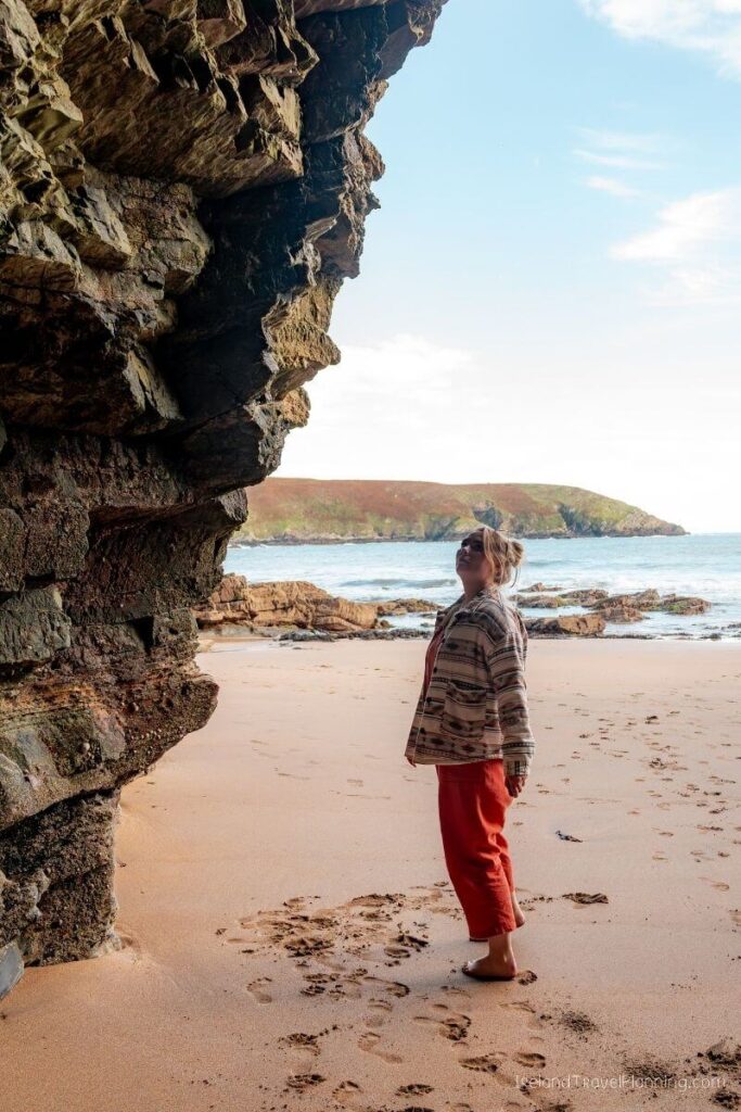 Woman in a patterned jacket and orange pants looking up at a rocky cliff face on a sandy beach.