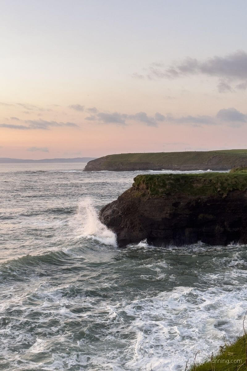 Waves crash against a rocky cliff on the Irish coast at sunset.