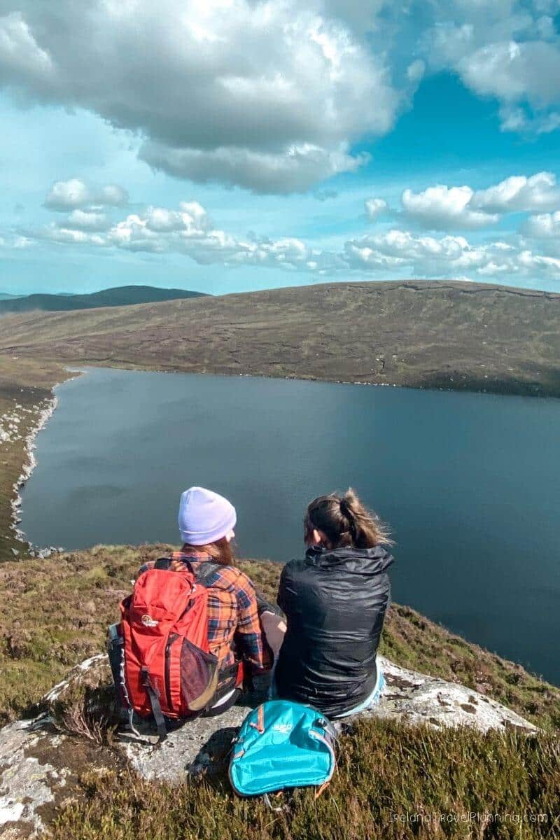 Two hikers admire a serene lake from a hilltop in Ireland.