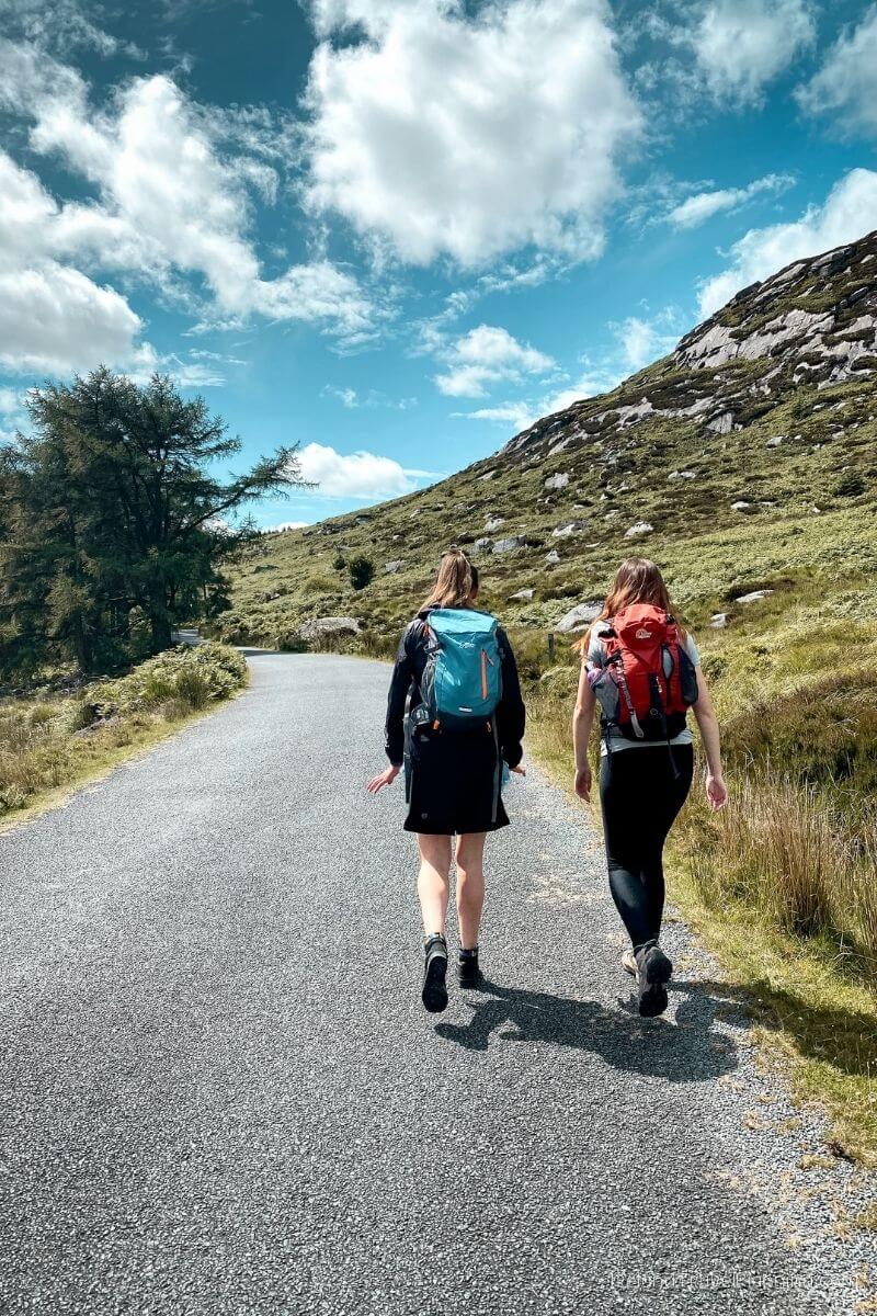 Two hikers with backpacks walk on a road in the Irish countryside.