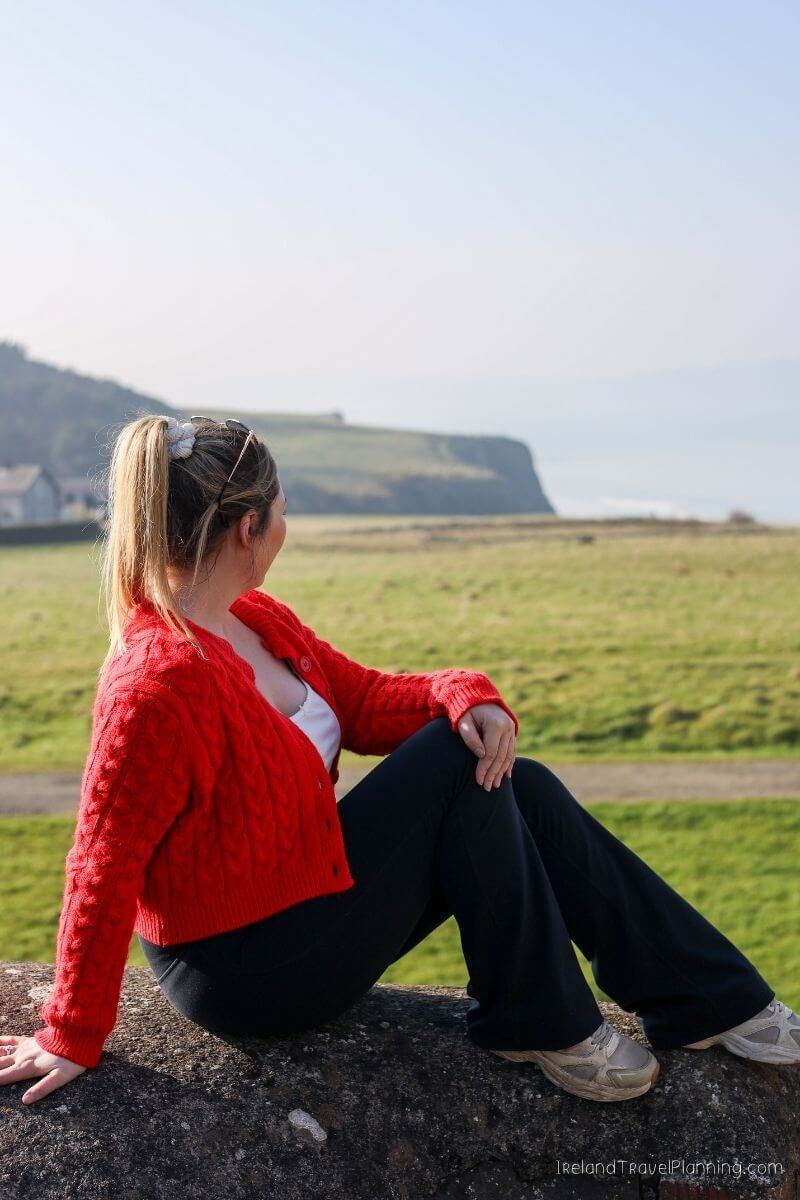 Woman in red cardigan overlooking Irish coastline, a hidden gem.