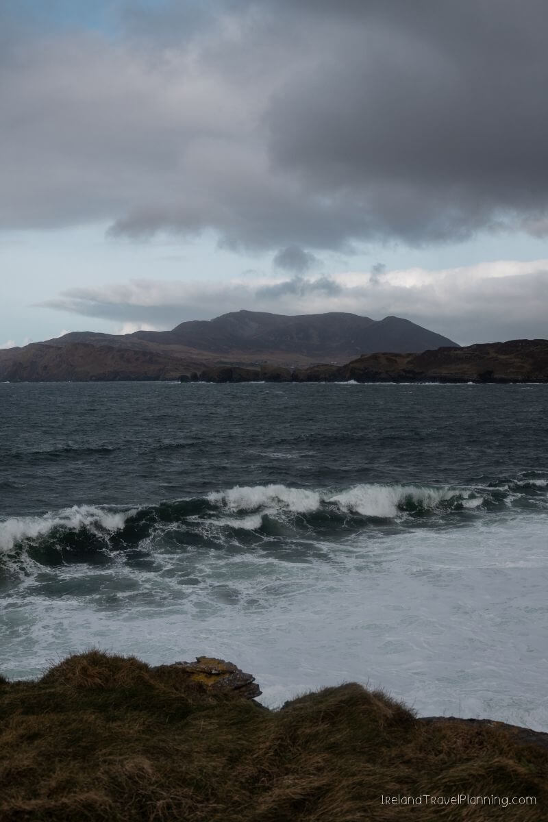 Dramatic waves crash on a rocky Irish coast with mountains in the background.