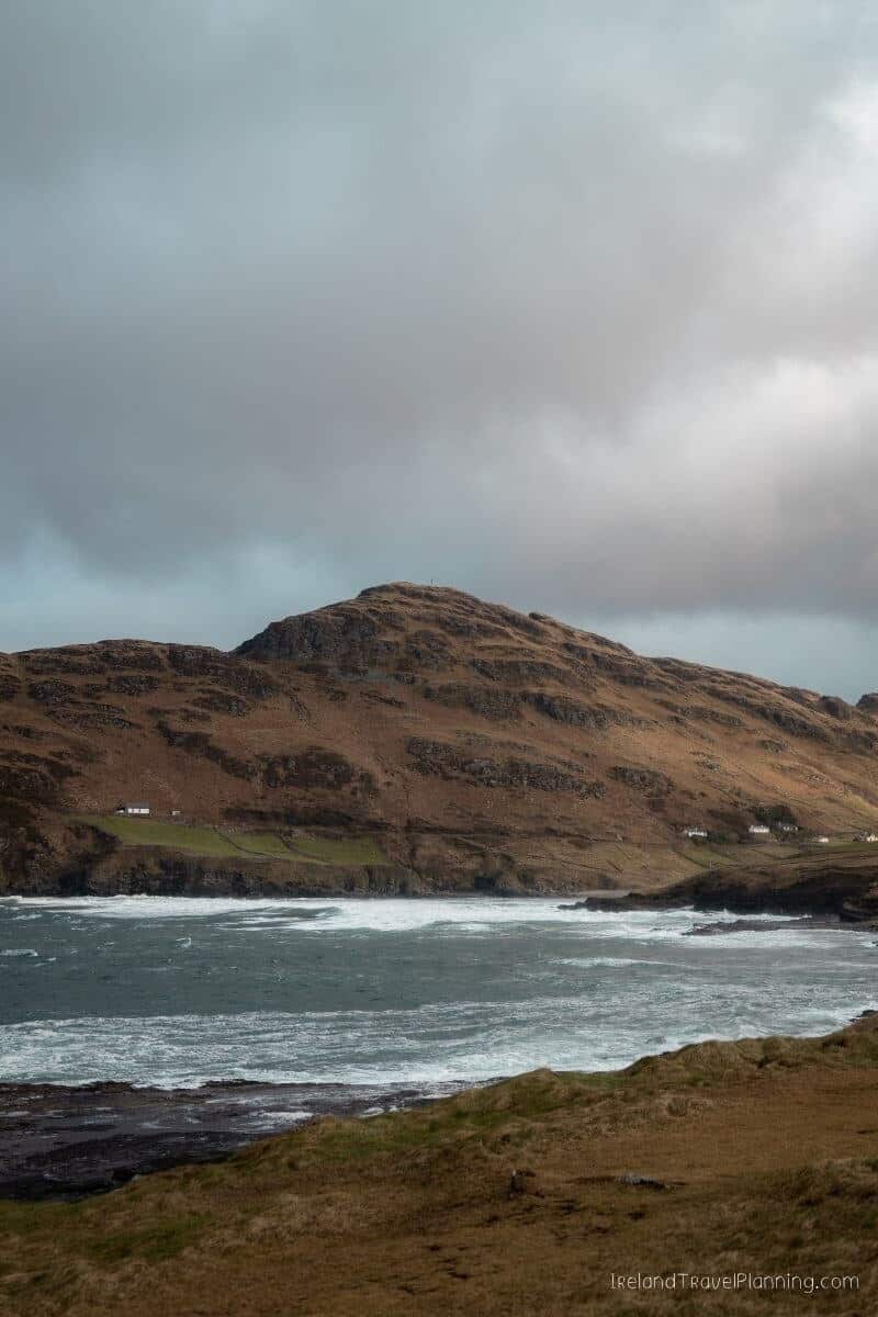 Rugged Irish coast with rolling hills, rough sea, and scattered white houses under a dramatic sky.