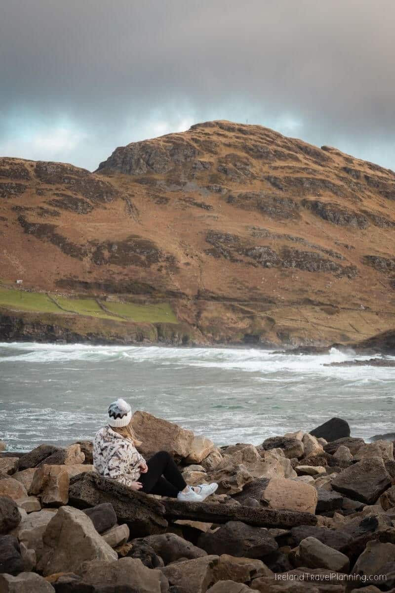 Woman in beanie and patterned jacket sits on rocks overlooking stormy Irish coast and rugged hill.