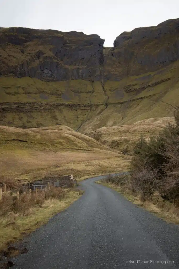 Winding asphalt road leading to a dramatic cliff face in a green, grassy valley.